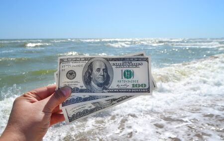 Girl holding a money bill of 300 dollars on background of sea waves and sky on sunny day. Hand waves sea ocean money dollars bills vacation relax summer. Concept finance money holiday horizon skylineの写真素材