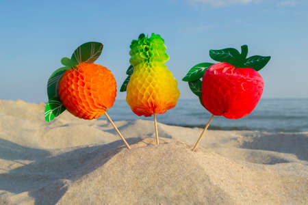 Three paper fruits on stick stand on sandy beach near sea on sunny summer day.の写真素材