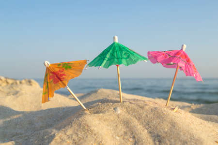 Three paper cocktail multi-colored umbrellas on sandy sea beach with blue skyの写真素材