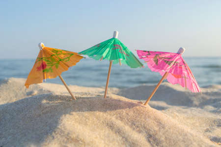 Three small paper cocktail umbrellas stand in sand on sandy beach close-up.の写真素材
