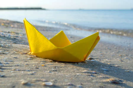 Small paper yellow boat on sand near water on background of sea wavesの写真素材