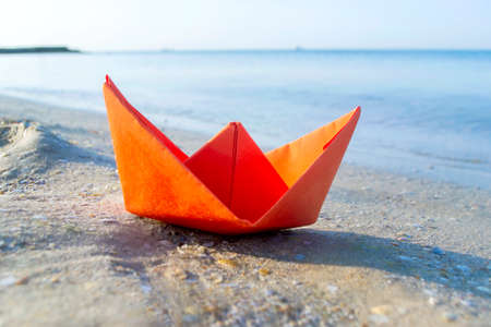 Small paper orange boat on sand near water on background of sea wavesの写真素材