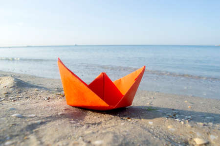 Small paper orange boat on sand near water on background of sea wavesの写真素材