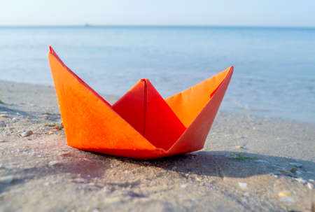 Small paper orange boat on sand near water on background of sea wavesの写真素材