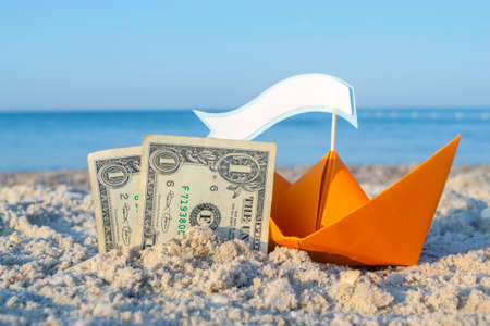 Two paper one dollars bills half buried in sand, orange paper boatの写真素材