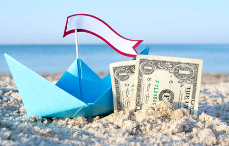 Two paper one dollars bills half buried in sand, blue paper boatの写真素材