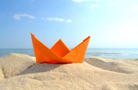 Bright orange paper boat in yellow sand on background of sea waves and blue skyの写真素材