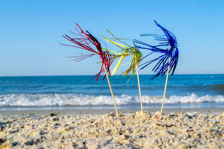 Three bright shiny colored cocktail decorations on stick stand in sand near seaの写真素材