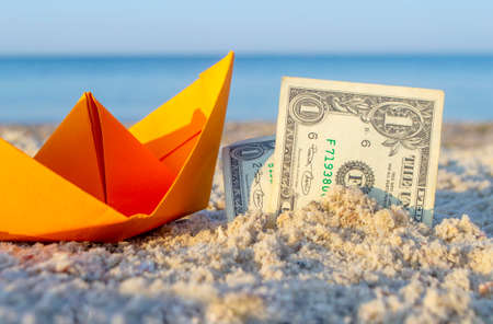 Orange paper boat and two paper dollar bills half buried in sand on sandy beachの写真素材