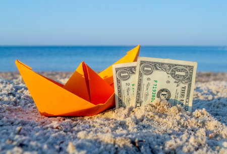 Orange paper boat and two paper dollar bills half buried in sand on sandy beachの写真素材