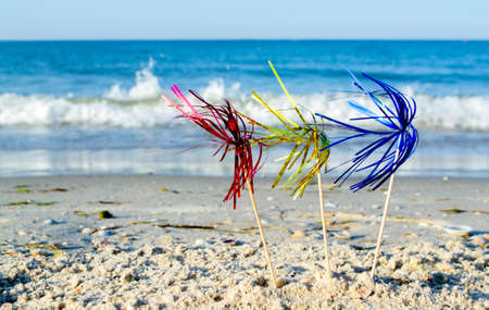 Three bright shiny colored cocktail decorations on stick stand in sand near seaの写真素材