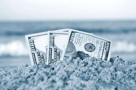 Three one hundred dollar bills half buried in sand on sandy seashore close-upの写真素材
