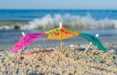 Three small paper cocktail umbrellas stand in sand on sandy beach close-up.の写真素材