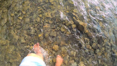 Girl walking barefoot on stones of shallow river close-up, sunny summer dayの写真素材