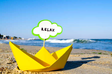 Small origami yellow paper boat with stick and speech bubble with word RELAX stands on sandy beach near sea water and sea waves with white foam close-up. Concept of travel, tourism, vacation, holidayの写真素材