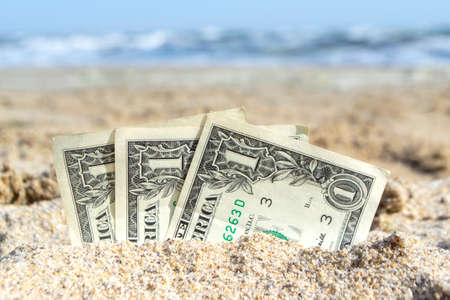 Three paper cash one dollar bills half buried in the sand on the seashore close-up. Concept money, business, savings, financial, investment, cash.の写真素材