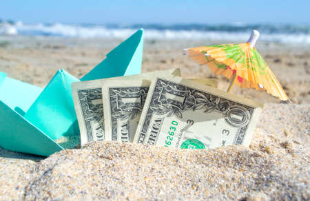 Blue small paper boat, half three dollar paper bills and paper cocktail umbrella in sand on sea beach on summer sunny day. Concept of travel dreams expenses money finance tourism rest vacation holidayの写真素材
