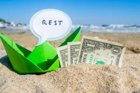 Small blue paper boat, three one paper dollar bills and speech bubble with word Rest on sandy sea...