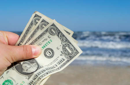 Person holding three paper dollar bills on background of sea and sea waves with white foam, sandy beach and clear blue sky on sunny summer day. Concept of money, expenses on vacation, travel, tourismの写真素材