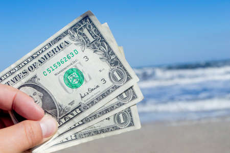 Person holding three paper dollar bills on background of sea and sea waves with white foam, sandy beach and clear blue sky on sunny summer day. Concept of money, expenses on vacation, travel, tourismの写真素材