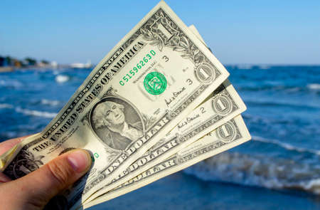 Person holds in his hand three dollar bills on the background of the waves of the blue sea. Concept money finance business tourism travel, money spending, cash expenses. Money cash expenses businessの写真素材