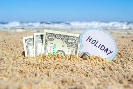 Paper bills one dollar and small stick with paper speech bubble with word Holiday buried in sand beach background sea close-up in sunny summer day. Concept Money travel tourism vacation holiday relaxの写真素材