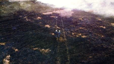 Two firefighters walk on black scorched earth after fire and burning dry grass in field. Firefighters walking on black land, ash after extinguishing fire. Aerial drone view. Red sun glare. Sun beamsの写真素材