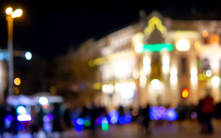 Blurred background. Blurred black silhouettes of people, blurred silhouette of architectural building illuminated by lanterns, backlight at night. City square in evening. Lots of people on city streetの写真素材