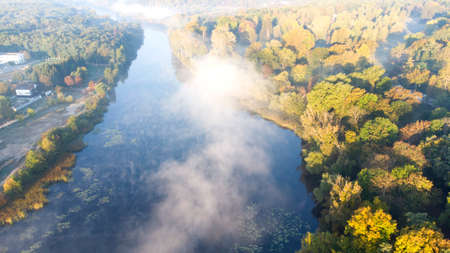 Aerial drone view flying over the river with calm reflective water surface and white fog mist and forest trees on the banks of the river on a sunny early autumn morning. Beautiful natural background.の写真素材