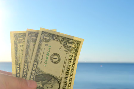 A man holding three one-dollar banknotes in his hand against backdrop of the clean blue sea and blue sky on sunny summer day. The concept of money, finance, monetary, travel, tourism, vacation, restの写真素材