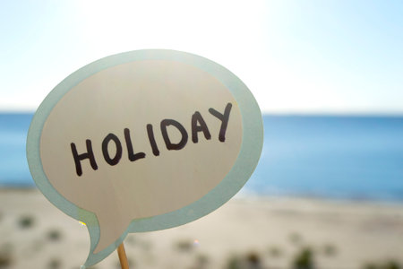 Stick with paper speech bubble with words Holiday on background blue sea, sky, sandy beach on sunny summer day. Text-balloons with text from letters. Concept, symbol, sign vacation travel tourism restの写真素材