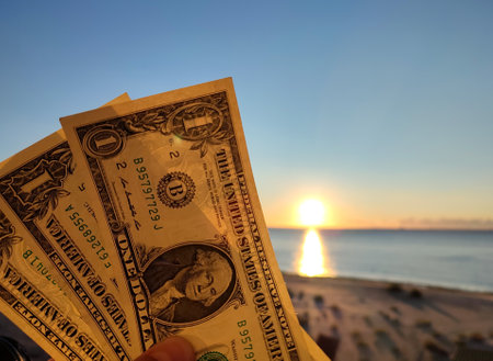 A man holding three one-dollar banknotes in his hand against backdrop of the sea, sandy beach and blue sky on sunny summer day. The concept of money, finance, monetary, travel, tourism, vacation, restの写真素材
