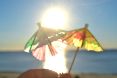 Man holding in hand three paper umbrellas cocktail decorations on background of blue sky, brightly shining sun and sunny path on surface of blue sea. Concept of summer, sea, vacation, travel, tourismの写真素材