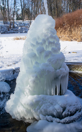 Iced fountain in the park on a winter sunny day. Fountain with frozen water. A jet of water from a fountain that iced over in the cold. Background covered with ice and snow in the park on dayの写真素材