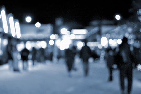 Blurred background. People walk in city square on winter night. Black silhouettes of people walking near houses decorated luminous illumination. White light bokeh spots glowing house lights Blue colorの写真素材