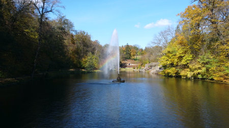 Fountain with rainbow in middle of lake between trees with yellow leaves in park on sunny autumn day. Decorative fountain in center of lake. Natural park arboretum in autumn. Natural landscape.の写真素材