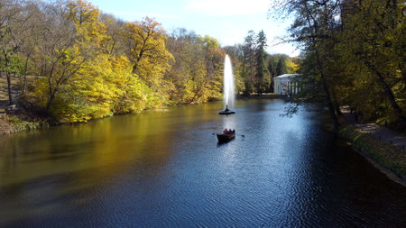 Beautiful landscape people sailing in boat on lake with decorative fountain among trees with yellow leaves in park on sunny autumn day. Natural background. Travel tourism rest recreation. Aerial viewの写真素材