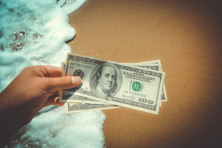 Girl holding money bill of 300 dollars on background of sea ocean waves with white foam and yellow sand wet beach close-up. Hand wave sea ocean money dollars. Concept finance money holiday travelingの写真素材