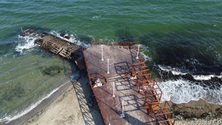 Old wooden pier with people resting and old concrete breakwater on the sandy beach of the sea shore on a sunny day. Public beach. Sea coast, sea shore. Coastal seascape. Top view. Aerial drone viewの写真素材