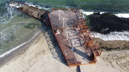 Old wooden pier with people resting and old concrete breakwater on the sandy beach of the sea shore on a sunny day. Public beach. Sea coast, sea shore. Coastal seascape. Top view. Aerial drone viewの写真素材