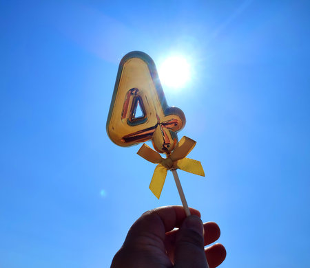 Person holding stick with a shiny golden number 4 four in his hand on backdrop of the waves of blue sky and shining sun on a sunny summer day. Concept holiday birthday party celebratingの写真素材
