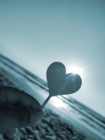 Person holding in fingers hand stick in shape heart on background sea and sea waves, blue sky in seashore on sunny summer day close-up. Concept love romance amour St Valentines Day. Blue colorの写真素材