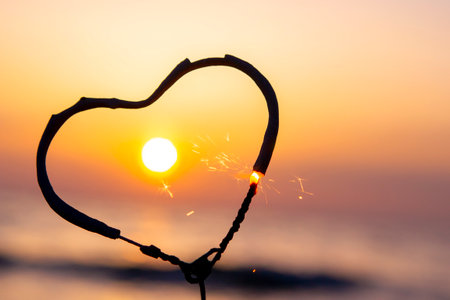 Heart-shaped sparkler burning against background of sea and rising sun at dawn. Bengal fire in shape of heart sparkling at sunrise dawn and sunset on sea. Love infatuation Valentines Dayの写真素材