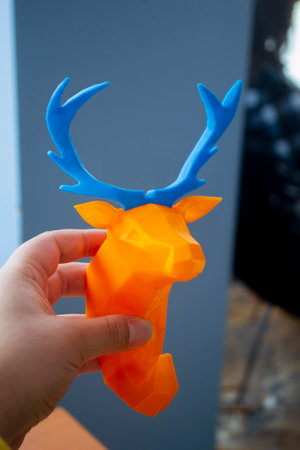 Person holding in hand an art object printed on 3D printer. Abstract toy model orange deer with blue...