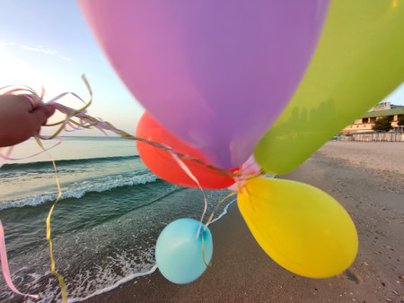 Hand and many colorful balloons tied with a ribbon near the sea on the sandy beach of seashore during sunrise and sunset. Multi-colored balloons at dawn. Seashore, sea coastの写真素材