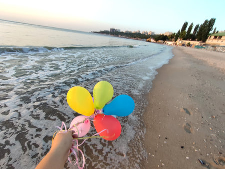Hand and many colorful balloons tied with a ribbon near the sea on the sandy beach of seashore during sunrise and sunset. Multi-colored balloons at dawn. Seashore, sea coastの写真素材