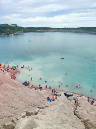 Many people swimming in the blue water near the sandy shore in a flooded quarry on a summer day. Beautiful blue water of large lake and many people swimming near the sandy shore of the slope. Top viewの写真素材