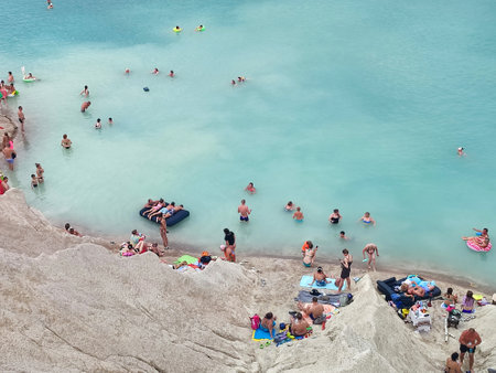Many people swimming in the blue water near the sandy shore in a flooded quarry on a summer day. Beautiful blue water of large lake and many people swimming near the sandy shore of the slope. Top viewの写真素材