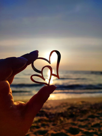 Woman holding in fingers black outline in shape of two hearts against background of sun at dawn and sunset on sea. Concept love infatuation romantic Valentines Day mood emotions relationshipの写真素材