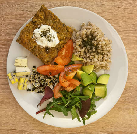 Cooked pearl barley, a piece of vegetable casserole with cream or natural yoghurt. Pieces of cheese with mold, tomato, avocado, arugula leaves, beetroot leaves, black lentil sproutsの写真素材